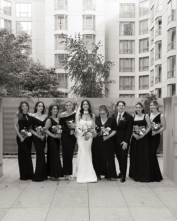 Wedding party portrait with bride and bridesmaids beside the groom in a black suit, holding bouquets on an outdoor patio by trees and a high-rise