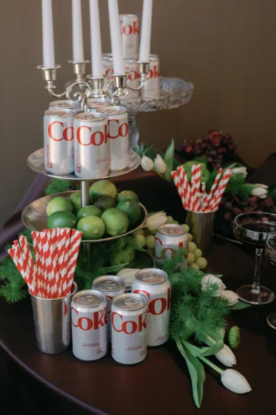 Wedding drink station with a wedding soda bar display of silver cans, red striped straws, limes, grapes, tulips, and lit candelabra on a dark tablecloth