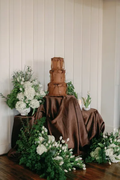 Wedding cake with ruffle frosting displayed on a brown satin tablecloth, framed by hydrangeas and tulips with pillar candles on pedestals