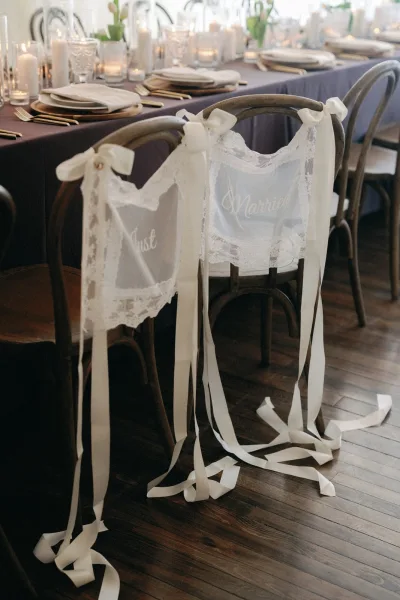 Wedding chair decor with bride and groom chair signs in lace and white ribbons, set at a reception table with gold flatware and candles
