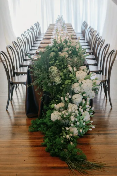 Reception tablescape with long banquet table wedding greenery garland, white hydrangeas and tulips, candles, and menus by bright windows