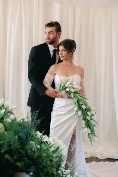 Couple portrait with bride in strapless lace wedding dress holding a cascading tulip bouquet as groom in black suit embraces her before white drapes