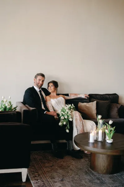 Couple portrait of bride and groom on sofa, bride holding white tulip bouquet, groom in tuxedo, with candles on coffee table in living room