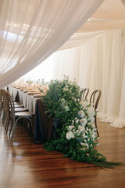 Reception tablescape with long head table decor, greenery and white tulips, candles in glass holders under draped tent ceiling