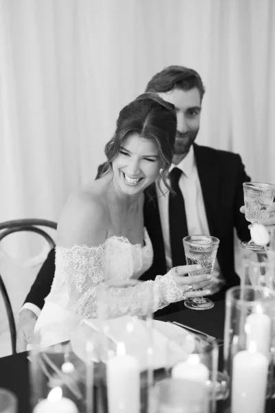 Wedding toast moment as bride and groom toasting with wine glasses at a candlelit reception table, white drapery backdrop and lace dress