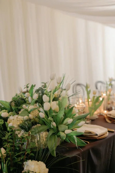 Reception tablescape with a tulip wedding centerpiece of white tulips and greenery, taper candles, and elegant place settings in a draped tent