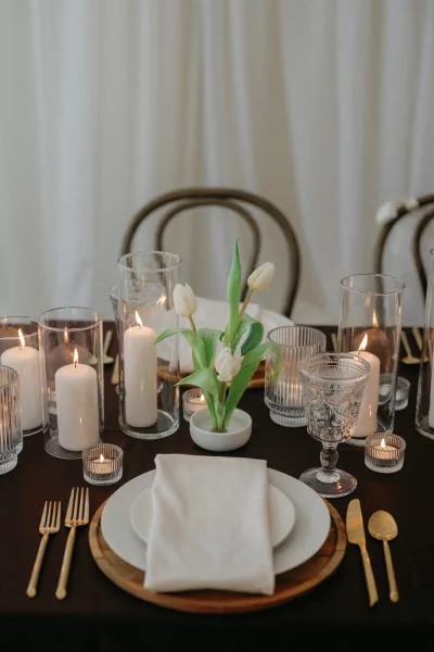 Reception tablescape with white tulip centerpiece and candlelit wedding table, gold flatware on wood chargers with bistro chairs and drapery backdrop