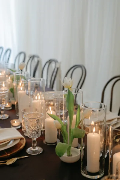 Reception tablescape with a candlelit wedding table of white pillar candles, tulips, vintage goblets, and wood runner under a draped tent wall