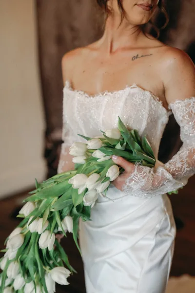 Bridal portrait of a bride holding bouquet of white tulips and greenery, wearing an off-shoulder lace gown in a neutral indoor room