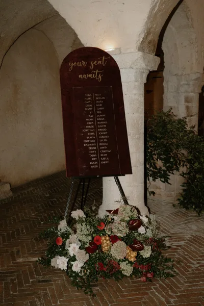 Wedding seating chart on an arched seating chart sign with calligraphy, on an easel beside roses and anthurium in a stone-arch courtyard