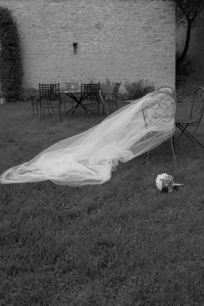 Bridal veil with cathedral veil train draped over a metal patio chair beside a bouquet and set outdoor table on a grassy lawn by a stone wall