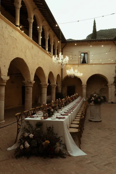 Reception tablescape with a long banquet table set with taper candles and floral garlands under crystal chandeliers in a stone-arched courtyard