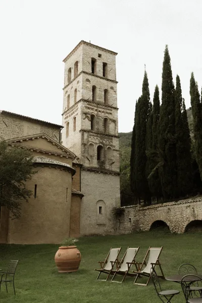 Wedding venue exterior with metal patio table and lounge chairs beside a terracotta urn, framed by a stone church tower and cypress trees