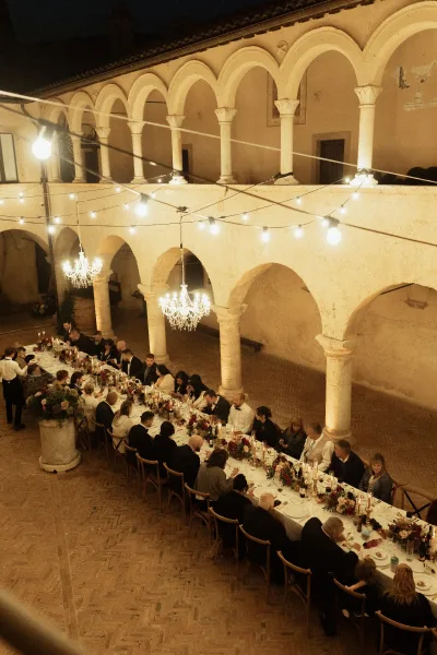 Wedding reception dinner at a long banquet table wedding with taper candles, burgundy florals, wine, and chandeliers under string lights in an arched courtyard