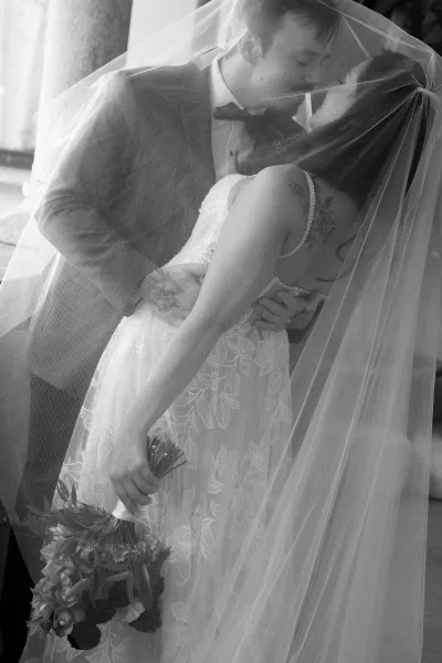 Wedding kiss portrait with a veil over couple kiss as the groom dips the bride in lace dress holding a bouquet in soft window light