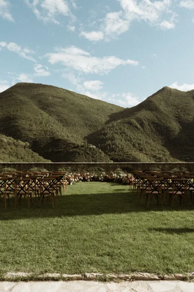 Ceremony setup with outdoor ceremony seating in rows of wood chairs, floral arrangements lining the aisle, and mountains beyond a stone wall under blue sky