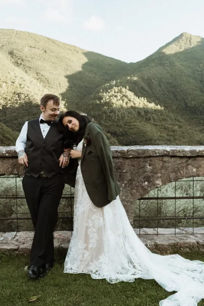 Couple portrait of bride leaning on groom in lace gown and green suit with bow tie, tattoos, and mountains behind a stone wall terrace