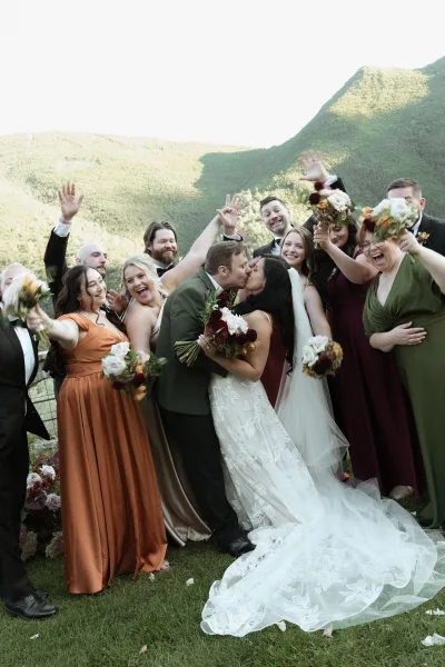 Wedding party photo of bride and groom kissing as friends cheer and raise bouquets on a mountain overlook with railing and sky backdrop