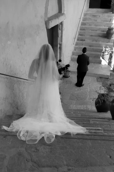 Wedding first look as bride walks up stone staircase toward groom in black suit, veil and dress train flowing, bouquet in hand by doorway