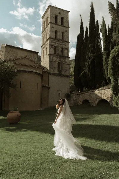Bridal portrait of a bride with long veil, holding a bouquet and looking back on a lawn before a stone church and cypress trees