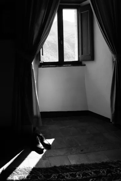 Groom shoes in black leather by a window with curtains, resting on a rug over tiled floor, with a mountain view outside