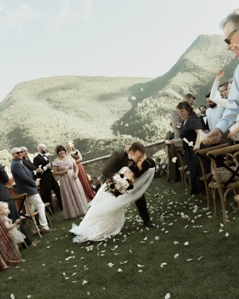 Wedding kiss as the groom dips the bride, veil flowing and bouquet in hand, rose petals flying down the aisle with mountain view behind guests