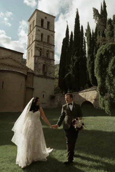 Couple portrait of bride and groom holding hands, her cathedral veil flowing by a stone church with bell tower and cypress trees