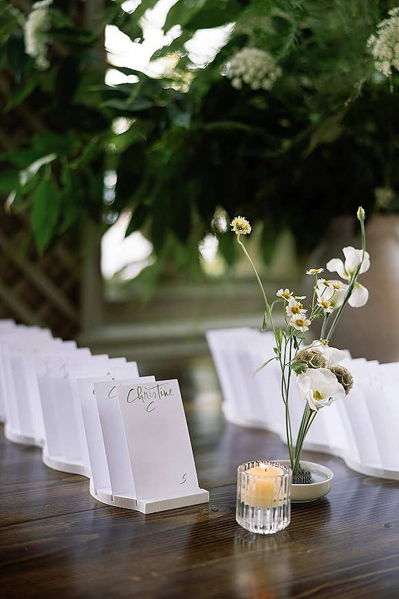 Wedding escort cards arranged in holders on a wooden table with a daisy bud vase and glass votive candle, softly lit by greenery and window light