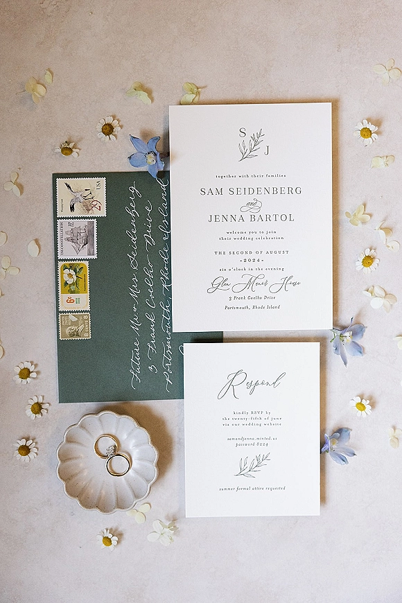 Invitation suite wedding invitation flat lay with RSVP card, calligraphy-addressed envelope, vintage stamps, rings, and blue daisies on light fabric backdrop