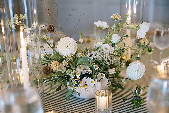Wedding centerpiece with a low floral centerpiece of white wildflowers and greenery, surrounded by taper candles and glass votives on a striped tablecloth