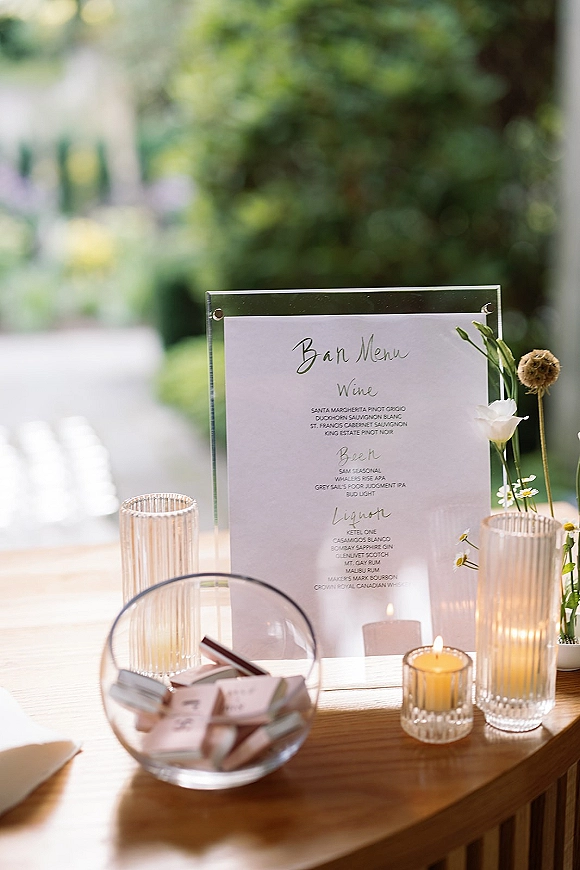 Wedding bar menu displayed in an acrylic holder with bud vases of white flowers and ribbed glass votive candles on a garden table backdrop