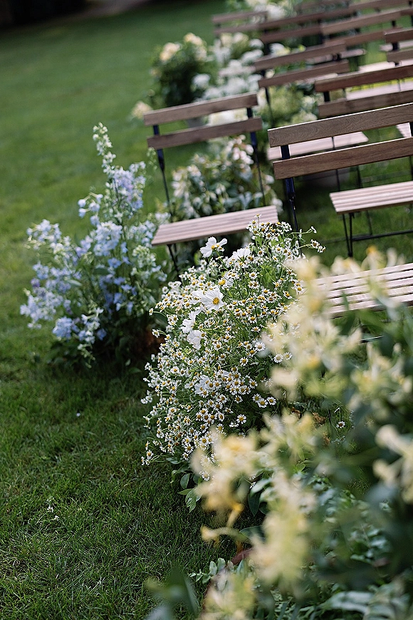 Ceremony aisle decor with outdoor wedding aisle flowers beside wood benches, featuring wildflower daisies and greenery on a garden lawn