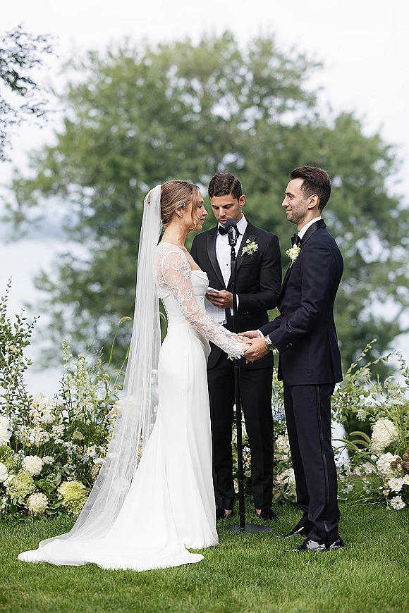 Wedding vows as bride and groom hold hands, her long veil and lace sleeves by a microphone stand on a garden lawn