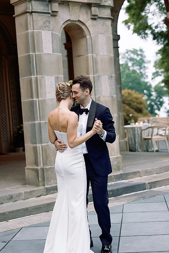 First dance moment outdoors as bride in a strapless wedding dress and groom in tuxedo touch foreheads beneath a stone archway