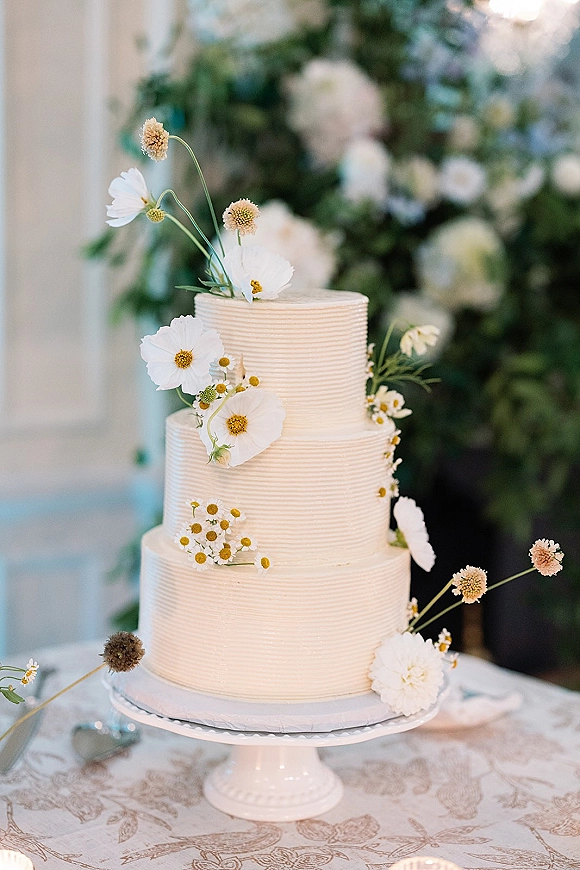 Wedding cake with white buttercream frosting, three tier design and cosmos and chamomile accents on a stand atop a lace tablecloth indoors
