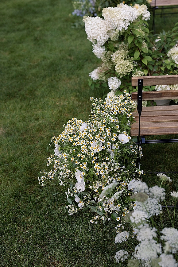 Garden wedding florals with white daisies and cosmos in a wildflower arrangement beside a wooden bench on a grassy lawn, hydrangea nearby