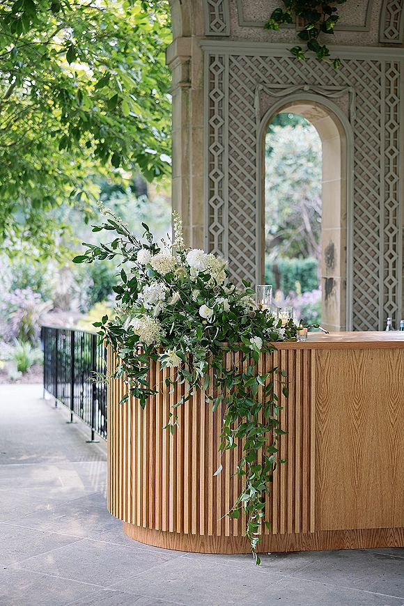 Wedding bar decor featuring a wood wedding bar with cascading white florals, ivy garland, and glass candle holders in a stone pavilion setting