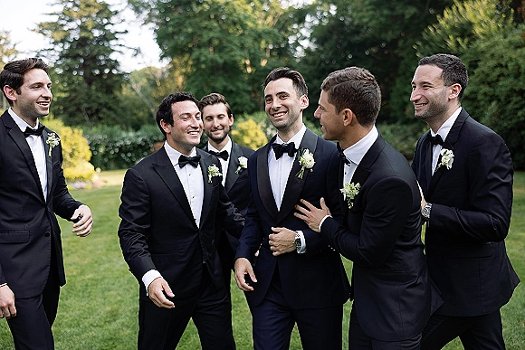Groomsmen portrait of men in black tuxedos and bow ties smiling together on a green lawn with trees in daylight