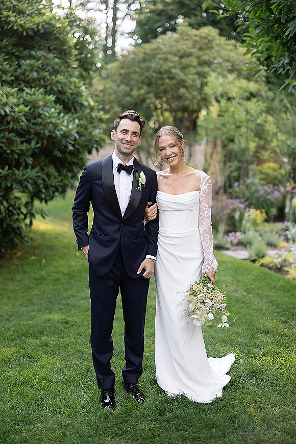 Couple portrait of bride and groom smiling arm in arm, her lace-sleeve wedding dress and bouquet beside his black tuxedo in a garden lawn setting
