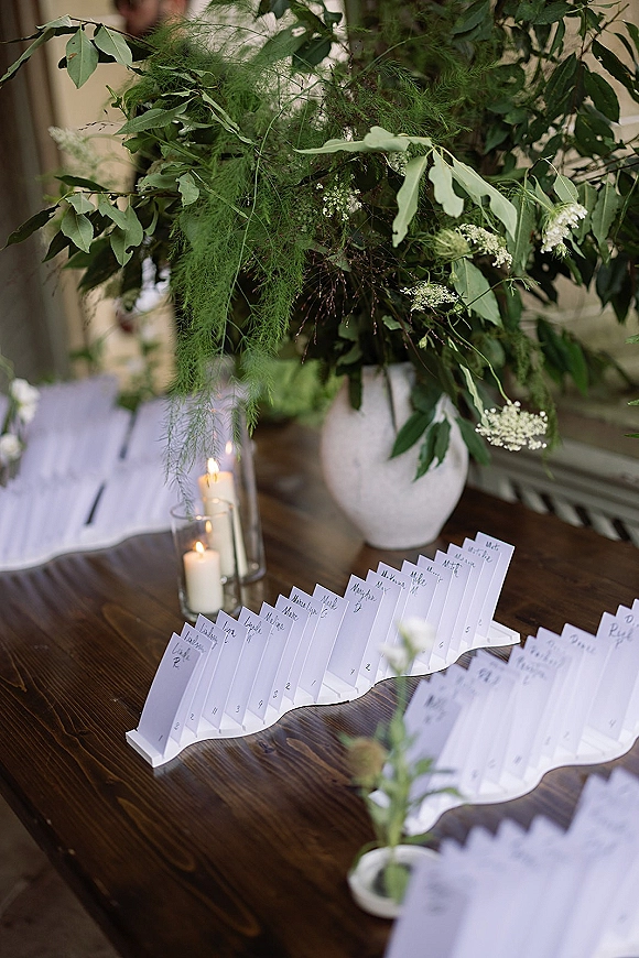 Wedding escort cards on an escort card table with wooden holders, a white vase and greenery centerpiece, and candlelight with guests blurred outdoors