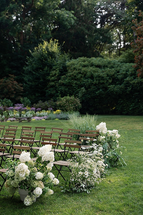 Ceremony setup with wood folding chairs and white hydrangea aisle flowers in pots along a grassy lawn bordered by trees and shrubs