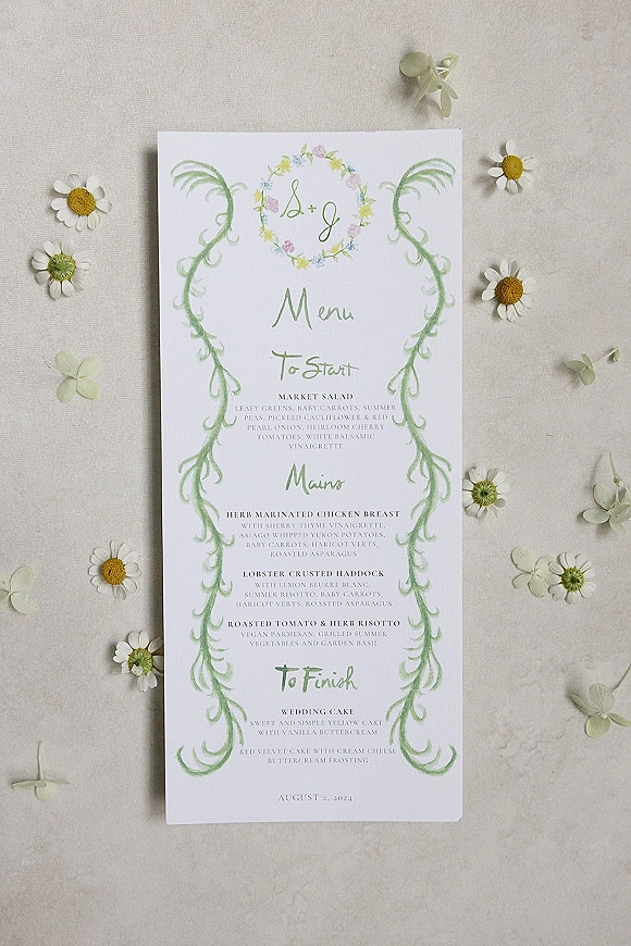 Wedding menu card with watercolor greenery and floral wreath monogram, printed typography and daisies, styled on linen fabric backdrop