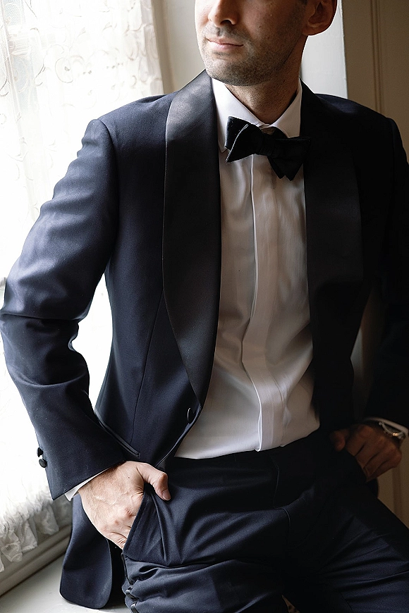 Groom portrait in a black tuxedo groom look with satin shawl lapel and bow tie, standing by window light with lace curtains indoors