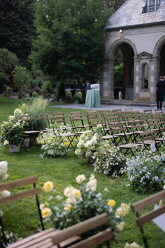 Ceremony seating setup with outdoor wedding ceremony chairs, wood folding chairs and blue-white aisle florals on a lawn by a stone arch doorway