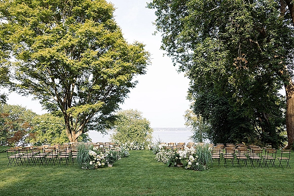 Outdoor ceremony setup with wood folding chairs and white floral aisle clusters on a lawn beneath large trees facing a lake view