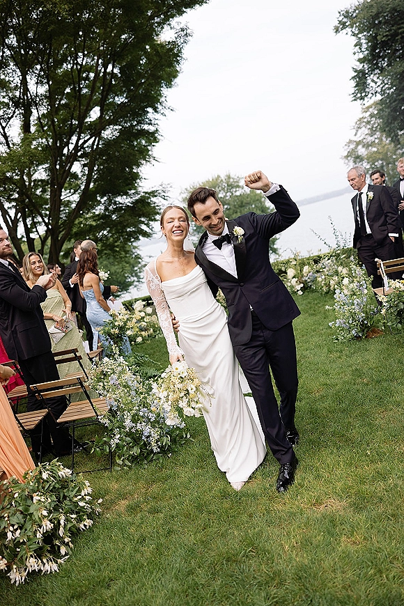 Wedding recessional as bride and groom walk the floral-lined aisle, groom fist-pumping while guests cheer at a lakefront lawn ceremony