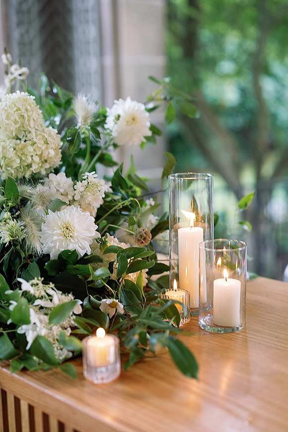 Wedding table decor with a wedding greenery garland, white hydrangeas and chrysanthemums, plus glass hurricane vases and pillar candles by windows with trees outside