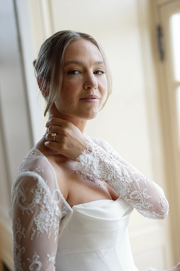 Bridal portrait of a bride by window light in a lace sleeve wedding dress, looking over her shoulder with updo and engagement ring
