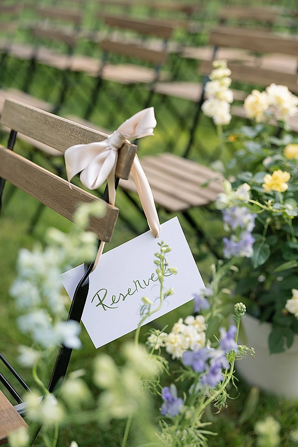 Reserved seating sign with wedding reserved sign calligraphy on a ribbon-tied satin bow over wooden folding chairs in a garden ceremony row