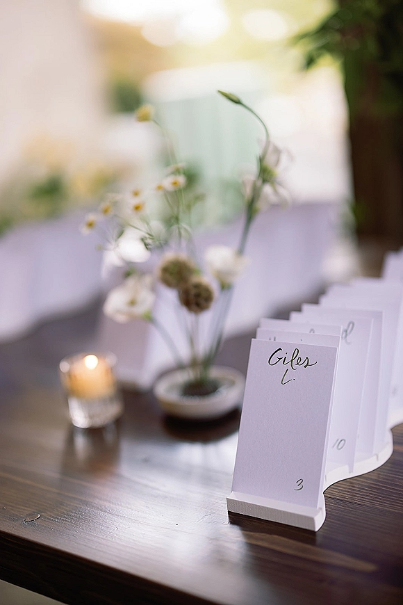 Wedding place cards with calligraphy place cards on holders beside a table number, bud vase of white flowers, and votive on a wood table in window light